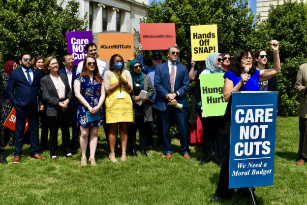 A group of people gather outside a government building with brightly colored signs that read "Hands off SNAP!", "Hunger Hurts," and "#HousingMatters." They stand behind a woman at a podium, with a sign "Care Not Cuts: We Need a Moral Budget." The woman at the podium is holding a mic and raising her fist.