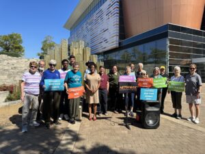 A group of advocates stand together behind a podium holding multi-colored signs that read, for example, "Protect Housing, "Protect WIC," "Protect Health Care." 