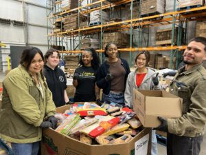 Coston fellows standing around a large bin of food that is being distributed to those in need.